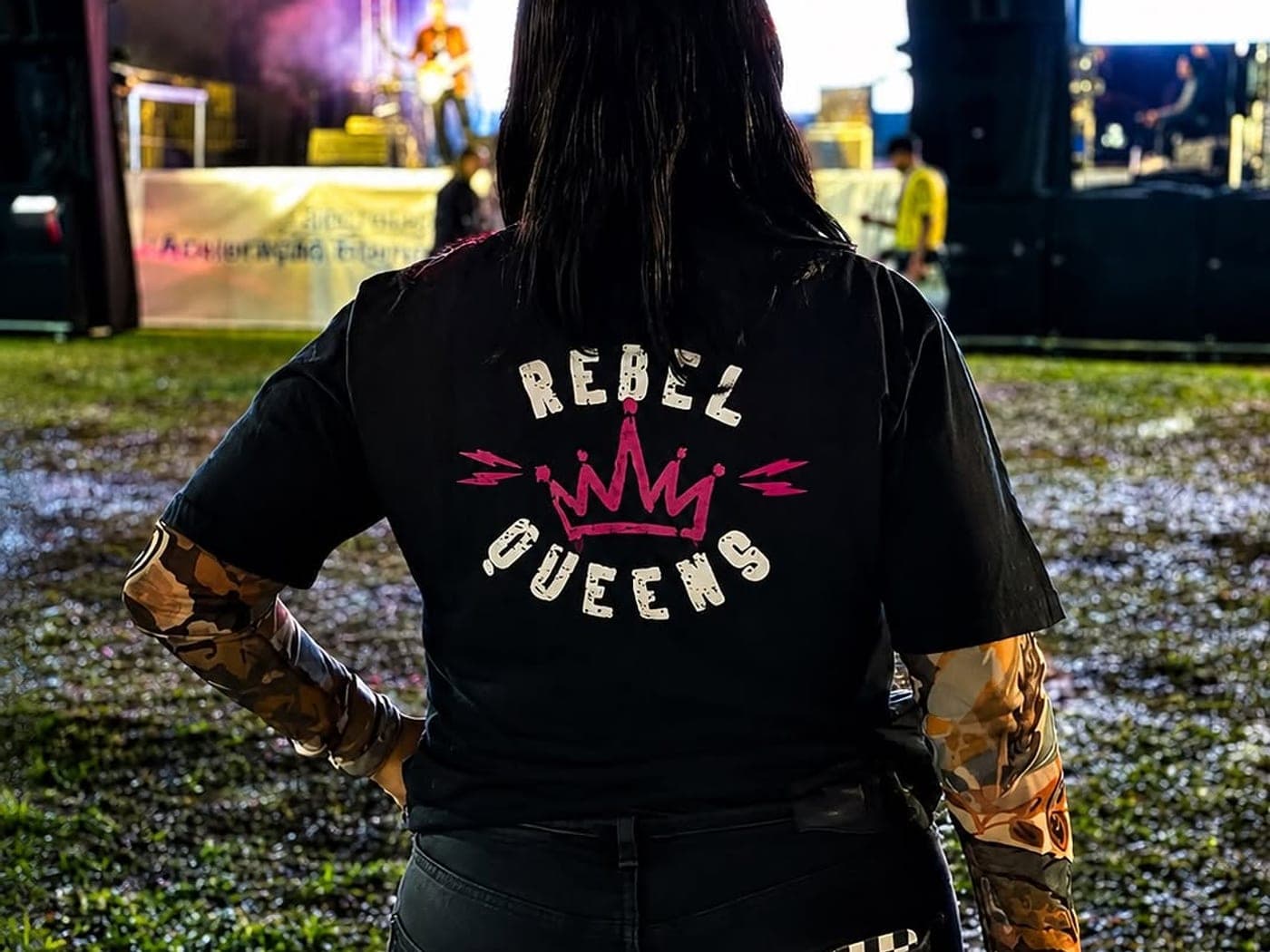 Motociclista de costas com a estampa Rebel Queens na camiseta, diante de um palco